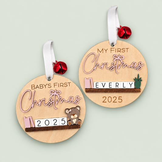 Two wooden baby's first Christmas ornaments with pink lettering, miniature shelf decorations and white ribbons on soft sage.