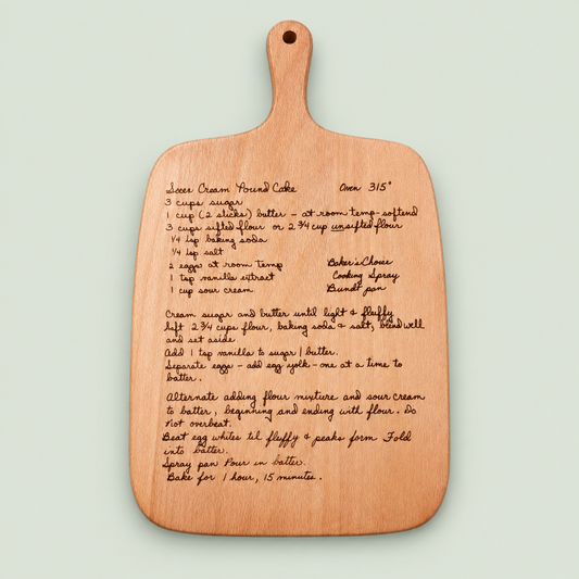 Wooden cutting board engraved with a handwritten sour cream pound cake recipe on a soft sage background.