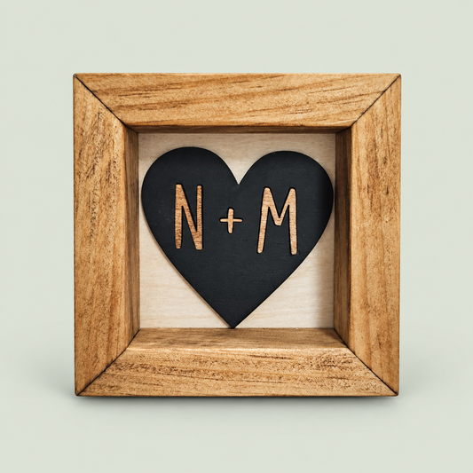 Wood-framed black heart sign with personalized N and M initials on a soft sage background.