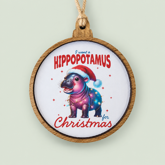 Wood-framed Christmas ornament featuring a colorful baby hippopotamus in a Santa hat on soft sage.
