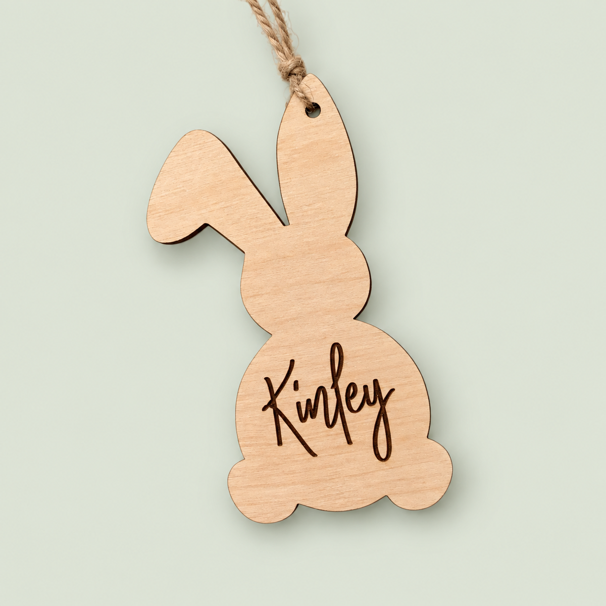 Wooden bunny Easter basket tag engraved Kinley with a twine hanger on a soft sage background.